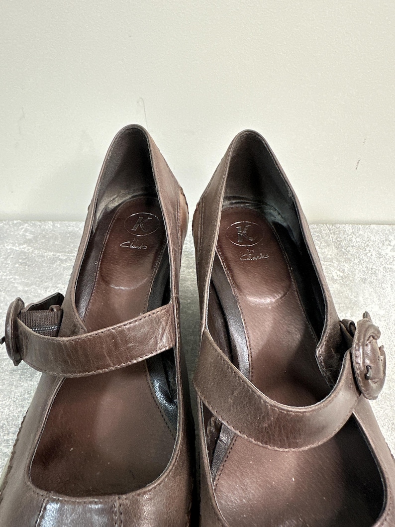 Może przedstawiać: Para brązowych sk&oacute;rzanych but&oacute;w na obcasie w stylu Mary Jane. Buty mają pasek na g&oacute;rze stopy z zapięciem na klamrę. Obcasy są średniej wysokości. Marka "Clarks" jest widoczna na wkładce.