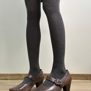 Może przedstawiać: Brązowe sk&oacute;rzane buty w stylu Mary Jane z zapięciem na klamrę i małym obcasem. Buty są noszone z szarymi rajstopami. Buty mają ozdobne przeszycia na krawędziach.