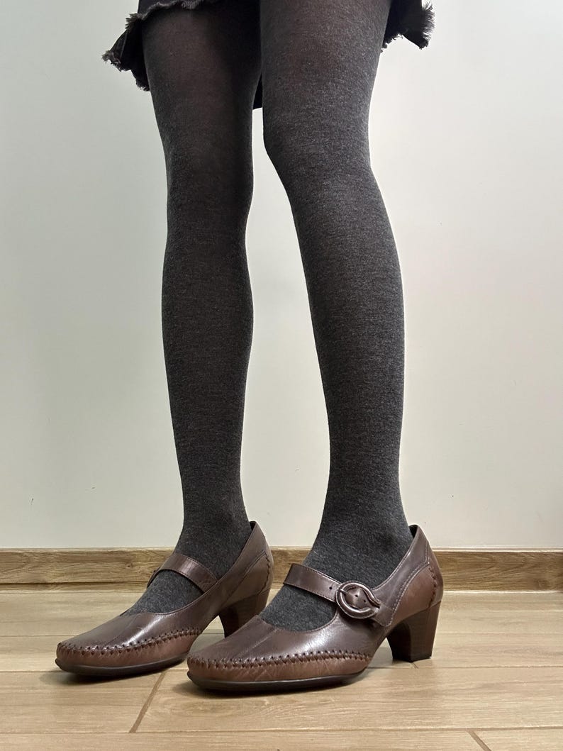 Może przedstawiać: Brązowe sk&oacute;rzane buty w stylu Mary Jane z zapięciem na klamrę i małym obcasem. Buty są noszone z szarymi rajstopami. Buty mają ozdobne przeszycia na krawędziach.