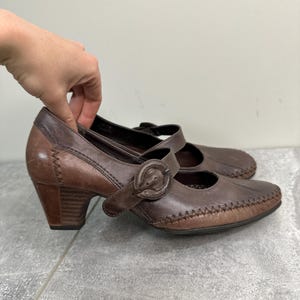 Może przedstawiać: Brązowe sk&oacute;rzane buty w stylu Mary Jane z zapięciem na klamrę i obcasem w słupku. Buty mają szyty wz&oacute;r wzdłuż krawędzi i zaokrąglony nosek. Wysokość obcasa wynosi około 6,35 cm.