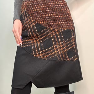 Op de afbeelding: Een stijlvolle rok met een patchwork-ontwerp. Het bovenste gedeelte is een bruin en oranje geweven stof, die overgaat in een zwart geruit patroon en eindigt met een effen zwart paneel aan de zoom. De rok is knielang.