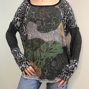 May include: A long-sleeved top with a unique patchwork design. The top features a black neckline and sleeves with a floral pattern. The center panel showcases a mix of green, gray, and brown floral and abstract patterns, creating a layered effect.