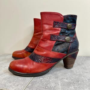 May include: A pair of red ankle boots with a stacked heel. The boots feature a patchwork design with blue and black accents, decorative buckles, and embossed leather details. The boots have a round toe and a sturdy heel.