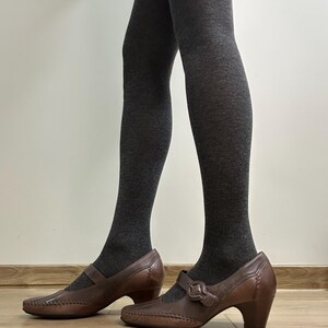 Może przedstawiać: Brązowe sk&oacute;rzane buty w stylu Mary Jane z zapięciem na sprzączkę i niskim obcasem. Buty mają ozdobne szwy i są noszone z ciemnoszarymi rajstopami. Buty stoją na drewnianej podłodze.