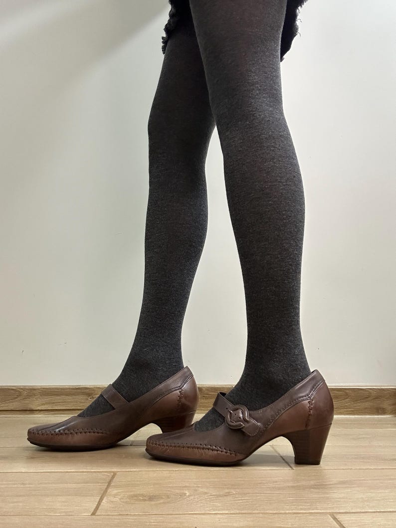 Może przedstawiać: Brązowe sk&oacute;rzane buty w stylu Mary Jane z zapięciem na sprzączkę i niskim obcasem. Buty mają ozdobne szwy i są noszone z ciemnoszarymi rajstopami. Buty stoją na drewnianej podłodze.