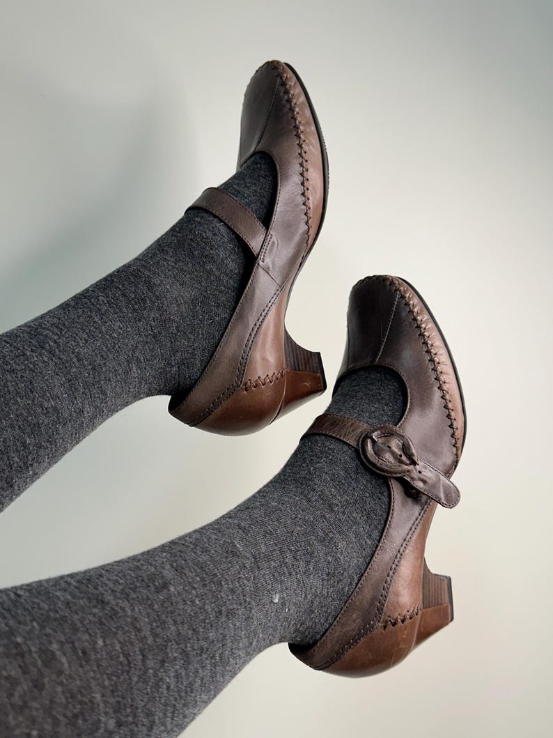 Może przedstawiać: Brązowe sk&oacute;rzane buty Mary Jane z paskiem i zapięciem na klamrę. Buty mają mały obcas i ozdobne szwy na krawędziach. Buty są noszone z szarymi skarpetami.