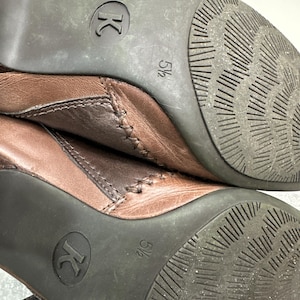 Może przedstawiać: Zbliżenie dw&oacute;ch brązowych sk&oacute;rzanych but&oacute;w. Podeszwy są ciemnobrązowe z teksturowanym wzorem i widocznym rozmiarem 5 1/2. Buty mają pasek i detale szw&oacute;w.