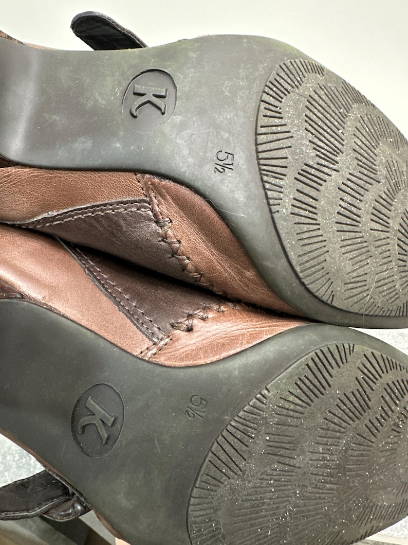 Może przedstawiać: Zbliżenie dw&oacute;ch brązowych sk&oacute;rzanych but&oacute;w. Podeszwy są ciemnobrązowe z teksturowanym wzorem i widocznym rozmiarem 5 1/2. Buty mają pasek i detale szw&oacute;w.