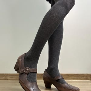 Może przedstawiać: Brązowe sk&oacute;rzane buty w stylu Mary Jane z zapięciem na klamrę i małym obcasem. Buty mają szyty wz&oacute;r na krawędziach. Buty są noszone z szarymi rajstopami.