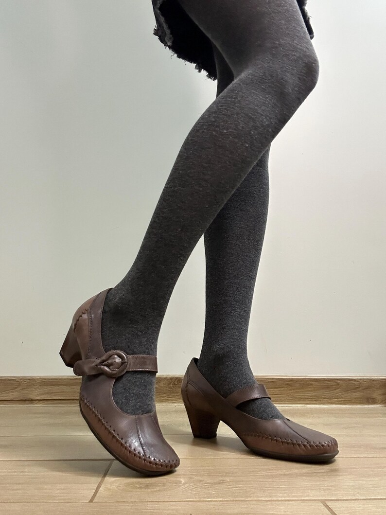 Może przedstawiać: Brązowe sk&oacute;rzane buty w stylu Mary Jane z zapięciem na klamrę i małym obcasem. Buty mają szyty wz&oacute;r na krawędziach. Buty są noszone z szarymi rajstopami.