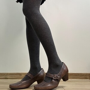 Może przedstawiać: Brązowe sk&oacute;rzane buty w stylu Mary Jane z zapięciem na klamrę i obcasem w kształcie słupka. Buty mają ozdobne przeszycia na palcach i cholewce. Osoba ma na sobie ciemnoszare rajstopy.