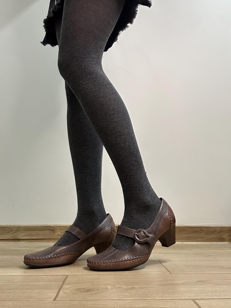 Może przedstawiać: Brązowe sk&oacute;rzane buty w stylu Mary Jane z zapięciem na klamrę i obcasem w kształcie słupka. Buty mają ozdobne przeszycia na palcach i cholewce. Osoba ma na sobie ciemnoszare rajstopy.
