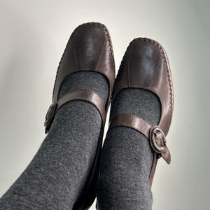 Może przedstawiać: Para brązowych sk&oacute;rzanych but&oacute;w w stylu Mary Jane z paskiem i zapięciem na klamrę. Buty mają zaokrąglony nosek i widoczne szwy. Buty są noszone z szarymi skarpetami.