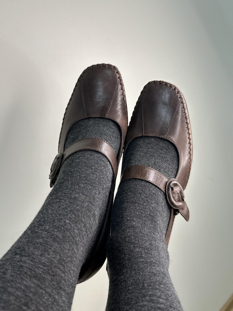 Może przedstawiać: Para brązowych sk&oacute;rzanych but&oacute;w w stylu Mary Jane z paskiem i zapięciem na klamrę. Buty mają zaokrąglony nosek i widoczne szwy. Buty są noszone z szarymi skarpetami.