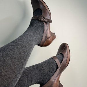 Może przedstawiać: Brązowe sk&oacute;rzane buty Mary Jane z paskiem i zapięciem na klamrę. Buty mają szyty wz&oacute;r na krawędziach i mały obcas. Buty są noszone z szarymi rajstopami.