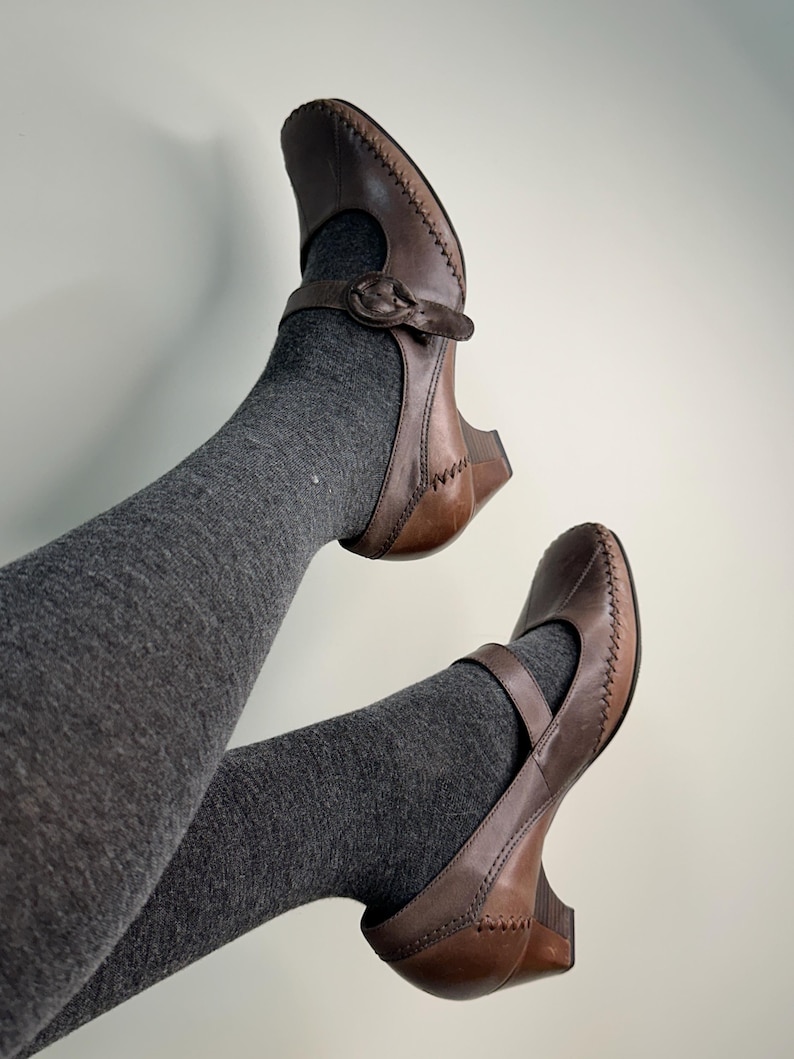 Może przedstawiać: Brązowe sk&oacute;rzane buty Mary Jane z paskiem i zapięciem na klamrę. Buty mają szyty wz&oacute;r na krawędziach i mały obcas. Buty są noszone z szarymi rajstopami.