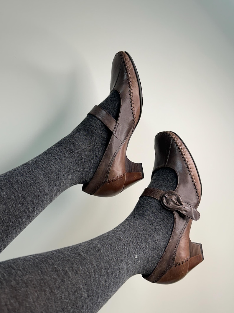 Może przedstawiać: Brązowe sk&oacute;rzane buty Mary Jane z zapięciem na klamrę i małym obcasem. Buty mają ozdobne przeszycia wzdłuż krawędzi i pasek na g&oacute;rze stopy. Buty są noszone z szarymi skarpetami.