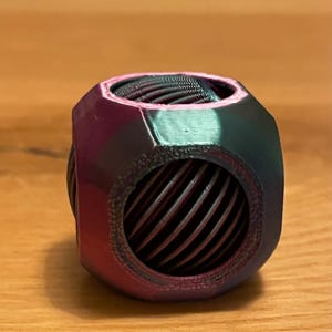 May include: A geometric object with a multi-faceted design and a circular aperture. The colour shifts from dark green to purple. The interior features a woven, metallic mesh. The design is modern and abstract.