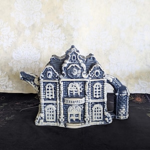 May include: A ceramic teapot shaped like a multi-story building with a dark blue and white color scheme. The structure features detailed architectural elements including arched windows, a central balcony, a clock face, and a textured roof.