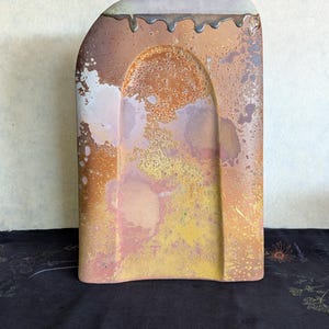 May include: A rectangular ceramic vase with a rounded top corner and a central arched indentation. The surface features a mottled glaze in shades of rust, ochre, yellow, and lavender, with a dark, drippy glaze accent along the top edge.