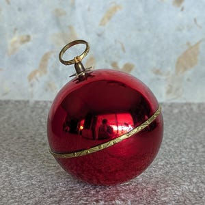 May include: A spherical red ornament with a reflective, glossy finish. The ornament features a thin, decorative gold-toned metal band encircling its center and a matching gold-toned metal loop at the top for hanging purposes.