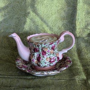 May include: A porcelain teapot and matching saucer set featuring a vibrant floral pattern of red and yellow roses on a green background. The teapot has a light pink spout and handle, with gold-toned trim along the scalloped edges of the rim.