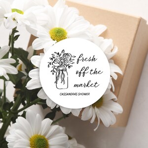 May include: A white circular sticker with the text "fresh off the market" and a floral illustration in black ink. The sticker is surrounded by white daisies with yellow centers and a brown box.