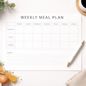 May include: A white weekly meal plan sheet with the text "WEEKLY MEAL PLAN" at the top. The sheet has columns for each day of the week and rows for breakfast, lunch, and dinner. A gold-tipped pen rests on the sheet.