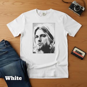 May include: White t-shirt featuring a black and white portrait of a musician. The image is a close-up of the musician's face. The word "White" is printed at the bottom of the image. A pair of blue jeans is next to the shirt.