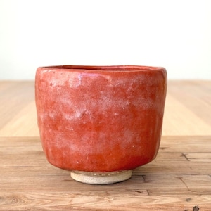 Może przedstawiać: Mała, cylindryczna ceramiczna miseczka z żywym, teksturowanym pomarańczowo-czerwonym szkliwem. Powierzchnia ma lekko nierówne, organiczne wykończenie z subtelnymi białymi akcentami. Podstawa miseczki jest nieszkliwiona, ukazując jasnobeżową, matową fakturę gliny.