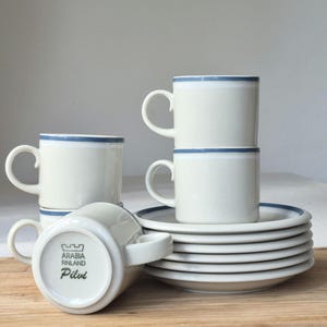 May include: A set of vintage Arabia Finland Pilvi coffee cups and saucers. The cups and saucers are off-white with a blue stripe around the rim. The cups are stacked and arranged with the saucers on a wooden surface.