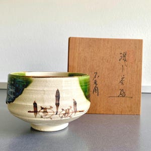 May include: A ceramic tea bowl with a cream-colored base, featuring a green glaze drip on the rim and brown painted motifs of vertical lines and circular shapes. Behind the bowl sits a square wooden storage box with black Japanese calligraphy.