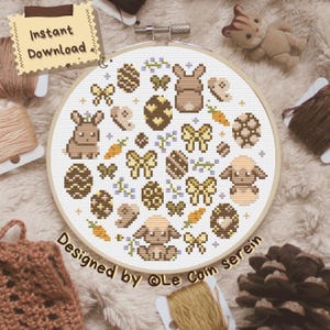 Peut inclure: Une broderie au point de croix dans un cercle en bois avec des motifs de Pâques. Le motif comprend des lapins bruns, des œufs décorés, des carottes et des nœuds. Le texte "Instant Download" et "Designed by @Le Coin Serein" sont visibles.