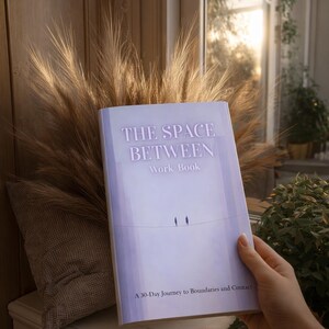 May include: A softcover book titled "THE SPACE BETWEEN Work-Book" with a minimalist design. The cover is light purple with the title in white and two small bird silhouettes. The book is held in front of a decorative arrangement.