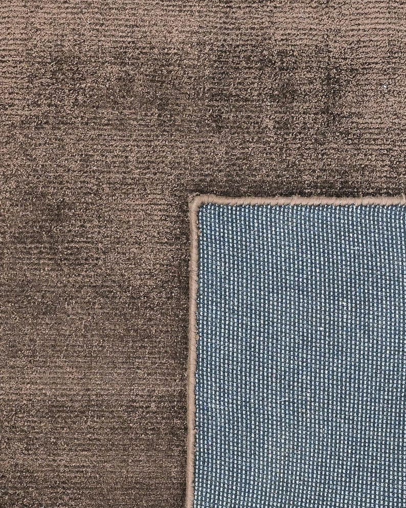 Pode incluir: Um close-up de um tapete marrom com uma superf&iacute;cie texturizada. O tapete tem uma borda listrada azul e branca. O tapete &eacute; feito de um material macio.