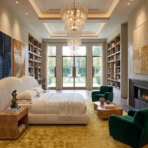 Puede incluir: Un dormitorio lujoso con una cama grande con cabecero color crema, ropa de cama blanca y una alfombra amarillo mostaza. Dos sillones verdes y una mesa de centro de madera est&aacute;n frente a una chimenea. Una gran l&aacute;mpara de ara&ntilde;a cuelga del techo.