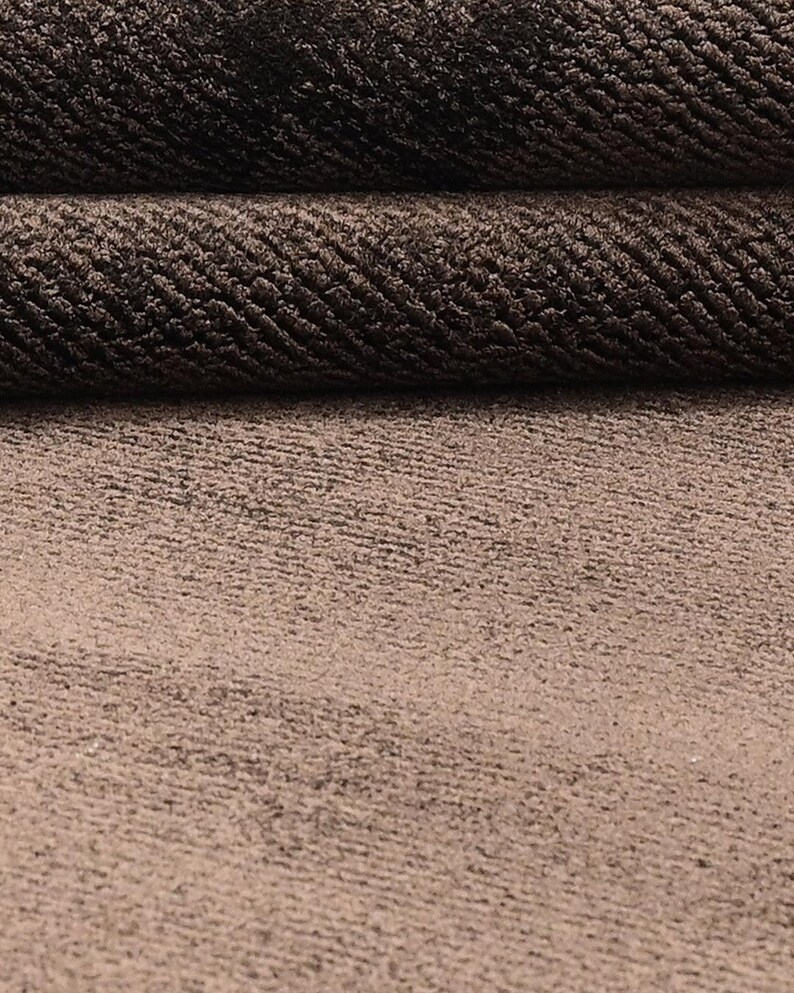 Pode incluir: Close-up de um tecido texturizado castanho. O material tem uma apar&ecirc;ncia tecida com um padr&atilde;o subtil. A imagem mostra o tecido dobrado sobre si mesmo, revelando sua textura e profundidade. Ideal para estofados ou projetos t&ecirc;xteis.