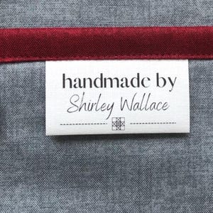 May include: A white label with the text "handmade by Shirley Wallace" in black. The label is on a gray fabric with a burgundy trim. A small geometric design is below the name.