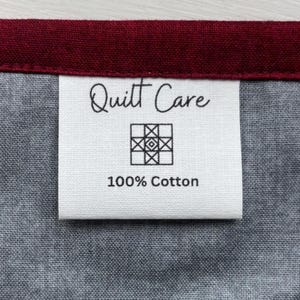 May include: A white rectangular fabric care label sewn onto a dark gray textile with a deep red border. The label features the black text Quilt Care, a small square geometric quilt block icon, and the text 100% Cotton printed in black ink.