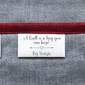 May include: A white rectangular fabric label featuring the black printed text A Quilt is a hug you can keep! with a small heart icon and the text By Sonja, stitched onto a grey textured fabric background with a horizontal dark red ribbon trim.