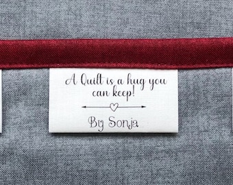May include: A white rectangular fabric label featuring the black printed text A Quilt is a hug you can keep! with a small heart icon and the text By Sonja, stitched onto a grey textured fabric background with a horizontal dark red ribbon trim.