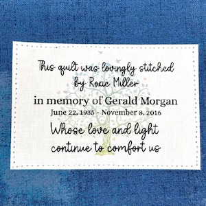 May include: A rectangular white fabric label with a stitched border featuring a printed tree graphic and black text that reads: This quilt was lovingly stitched by Roxie Miller in memory of Gerald Morgan June 22, 1935 - November 8, 2016 Whose love and light continue to comfort us.