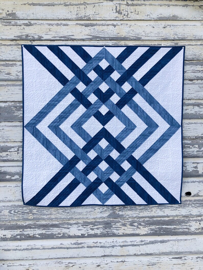 May include: A blue and white quilt with a geometric pattern. The quilt is made of different shades of blue fabric and is hanging on a white wooden wall.
