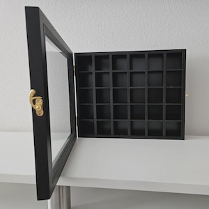 May include: Black display case with a glass door and a grid of small square compartments. The case has a gold-colored clasp and hinges. The open door reveals the interior, designed for displaying small collectibles.