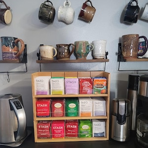 May include: A three-tiered wooden tea bag organizer filled with various tea brands including Tazo, Stash, and Twinings. The organizer sits on a counter next to a silver electric kettle and a coffee grinder, with ceramic mugs hanging above.