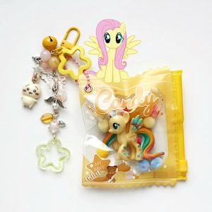 Op de afbeelding: Een gele sleutelhanger met een ster- en belbedel, en een doorzichtige plastic zak met een speelgoedpony erin. Op de zak staat het woord "Candy". Een cartoonpony met gele vacht en roze haar is aan de zak bevestigd.