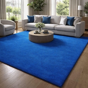 May include: A vibrant blue rectangular area rug dominates a living room scene. The rug is placed on a hardwood floor, with a light gray sofa, accent chairs, and a round coffee table. Decorative pillows and flowers add to the room's decor.