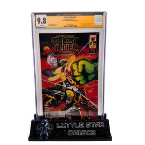 Op de afbeelding: Een beoordeeld Ghost Rider #4 stripboek, ingesloten in een beschermende houder, met een beoordeling van 9,8. De strip wordt weergegeven op een zwarte standaard met de tekst "LITTLE STAR COMICS". De cover bevat actievolle illustraties in rood, geel en groen.