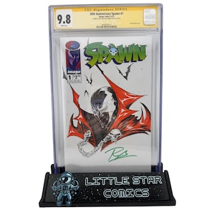 以下が含まれることがあります： CGCシグネチャーシリーズのコミック、Spawn #1、9.8点。表紙には、赤いマントとスカルチェーンを持つSpawnの詳細なイラストが描かれています。コミックは、「LITTLE STAR COMICS」の文字が入った黒いスタンドに保護ケースに入れて展示されています。