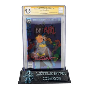 Peut inclure: Une bande dessinée notée mettant en vedette Batgirl, présentée dans un étui de protection. La couverture présente Batgirl avec d'autres personnages. La bande dessinée est notée 9,8 par CGC. Le présentoir indique "LITTLE STAR COMICS".